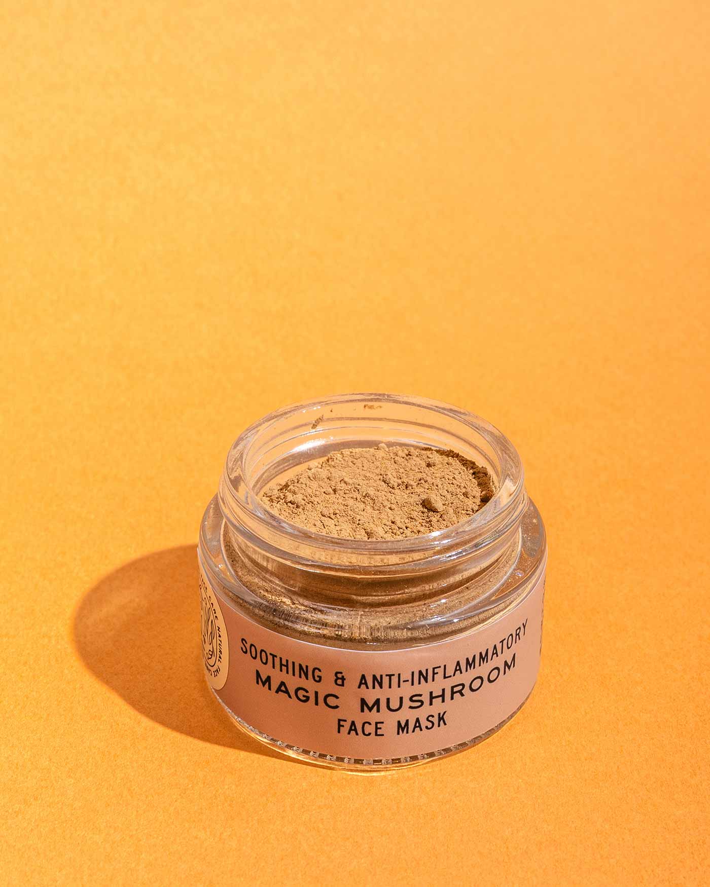 A clear jar labeled Magic Mushroom Face Mask by Good Flower Farm sits on an orange surface, filled with light brown rhassoul clay powder for a collagen-boosting glow.