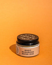 A small Good Flower Farm Magic Mushroom Moisture Cream jar with a black lid and beige label sits on an orange background. The cream is infused with adaptogenic maitake.