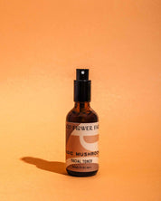 A brown glass spray bottle of Good Flower Farms Magic Mushroom Facial Toner stands upright on an orange surface, providing elemental protection and skin hydration, with a soft shadow beside it.