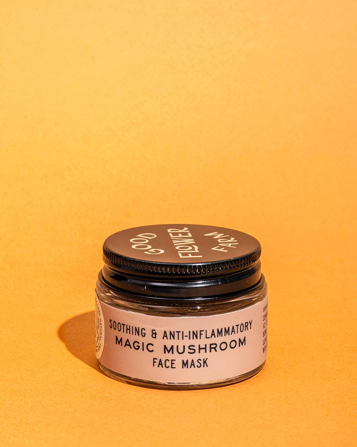 A small brown jar of Good Flower Farm’s Magic Mushroom Face Mask sits on an orange surface with an orange background. The collagen-boosting mask features a black lid with white text and a label that reads “Soothing & Anti-Inflammatory.”.