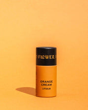A cylindrical orange container of Good Flower Farms TESTER — Orange Cream Organic Lip Balm stands upright against an orange background, casting a small shadow to the left. Its top is black with gold text.