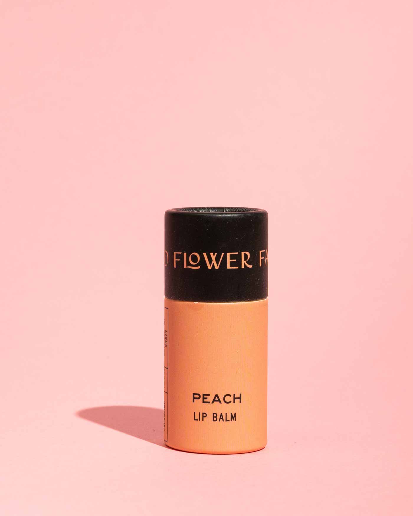 A moisturizing, peach-colored tube of Good Flower Farm TESTER — Peach Organic Lip Balm stands upright on a light pink background with a subtle shadow. The black cap features partially visible gold lettering.