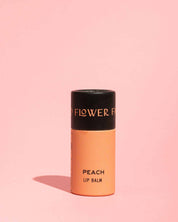 A moisturizing, peach-colored tube of Good Flower Farm TESTER — Peach Organic Lip Balm stands upright on a light pink background with a subtle shadow. The black cap features partially visible gold lettering.