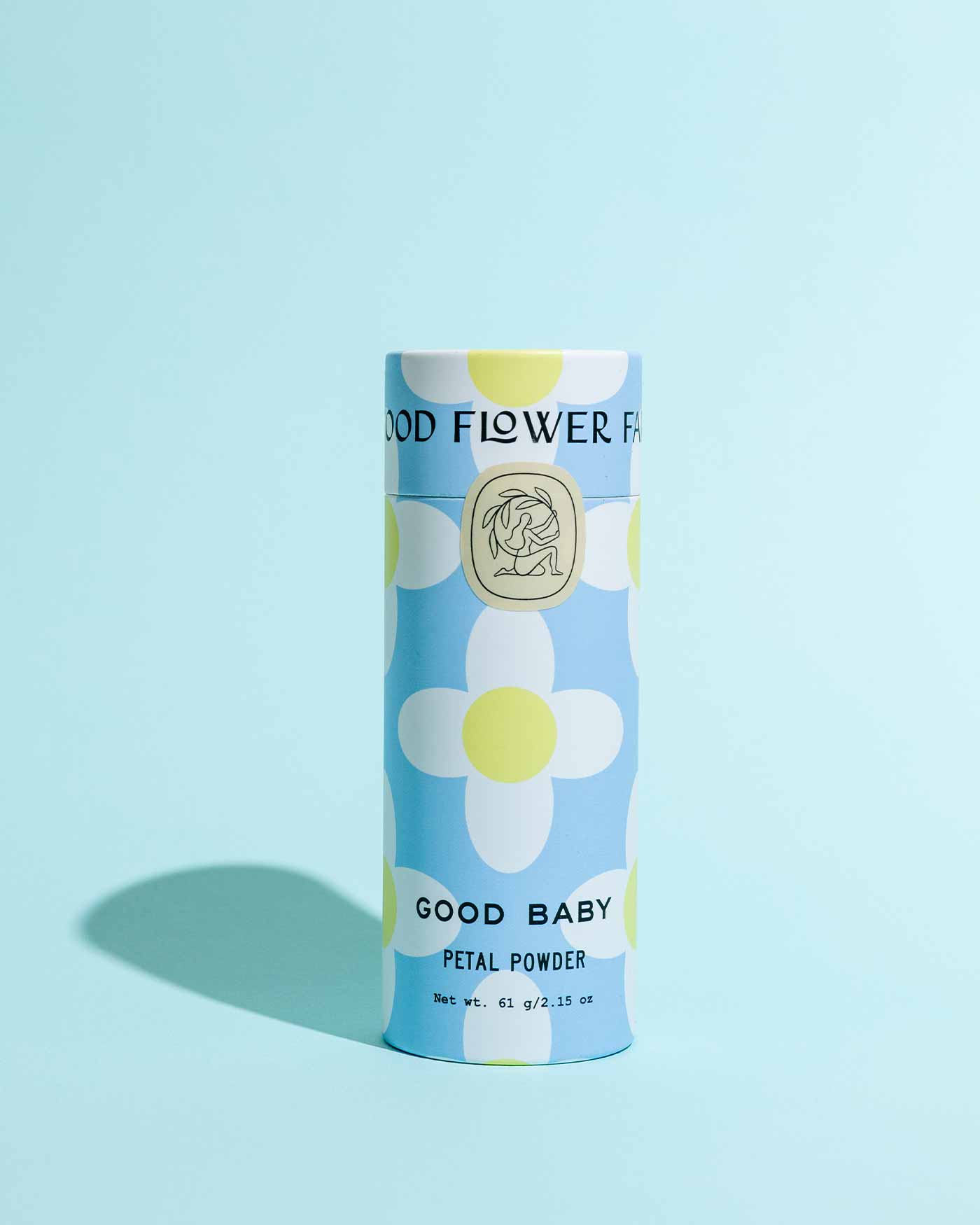 A cylindrical container of TESTER — Good Baby Petal Powder by Good Flower Farm stands upright on a light blue background, adorned with large white and yellow flowers, and features biodegradable, eco-friendly packaging.