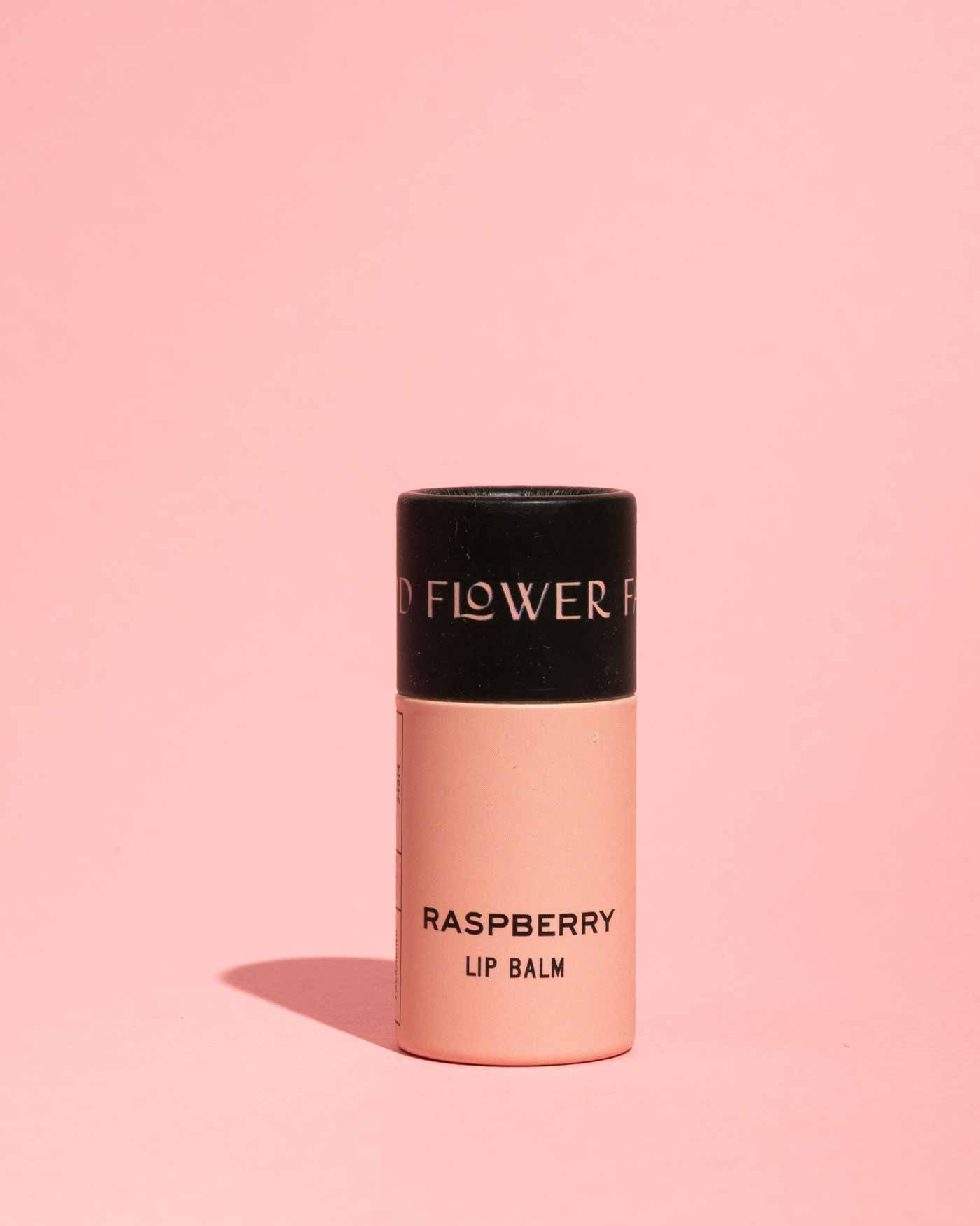 A tube of Good Flower Farm’s TESTER — Raspberry Organic Lip Balm, featuring a black cap and pink body labeled RASPBERRY LIP BALM, stands upright on a pink background with its shadow to the left—ideal for eco-friendly lip care.