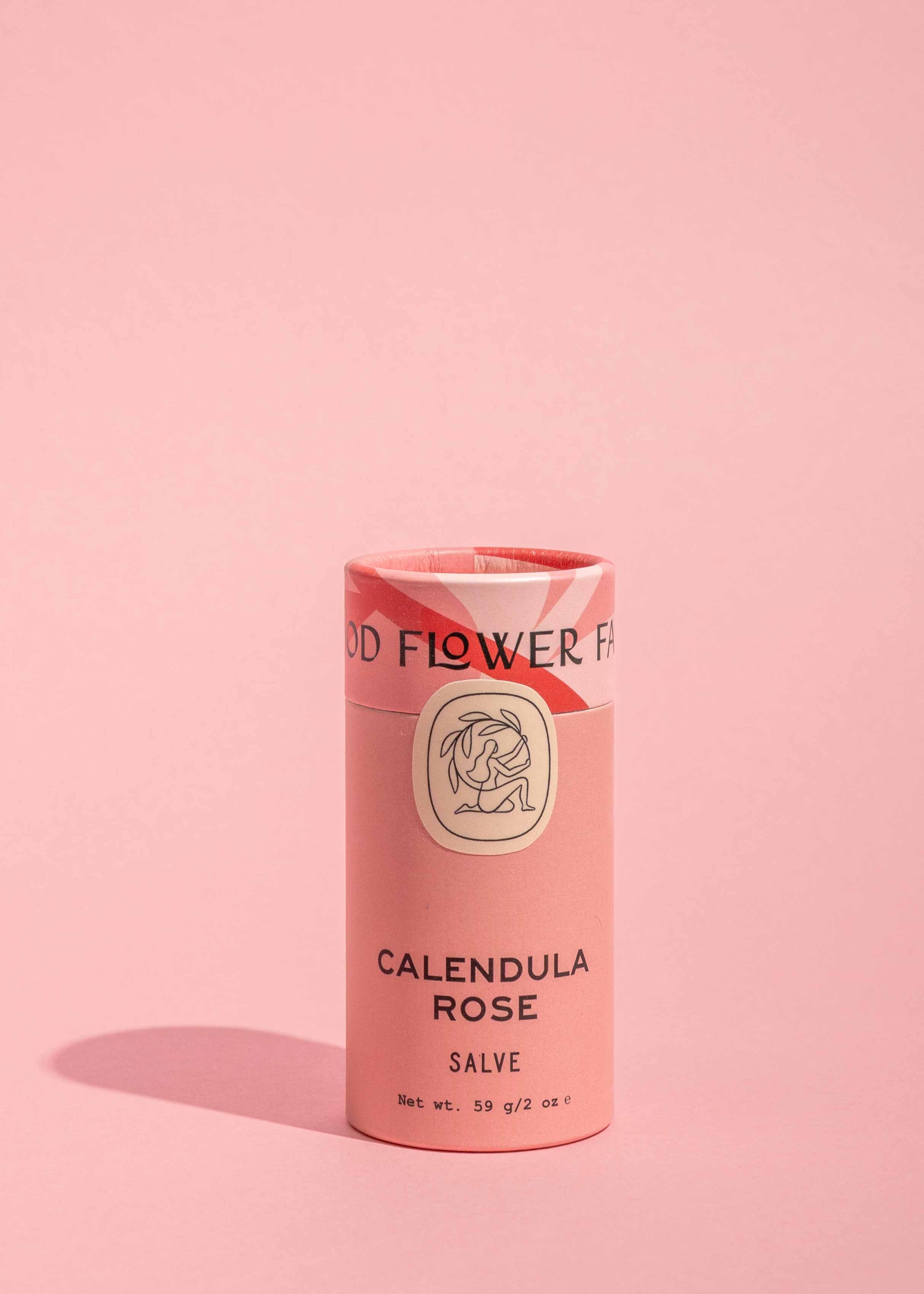A pink cylindrical container of Good Flower Farm Calendula Rose Salve stands upright on a matching background, featuring simple black text, a floral illustration, and eco-friendly packaging.