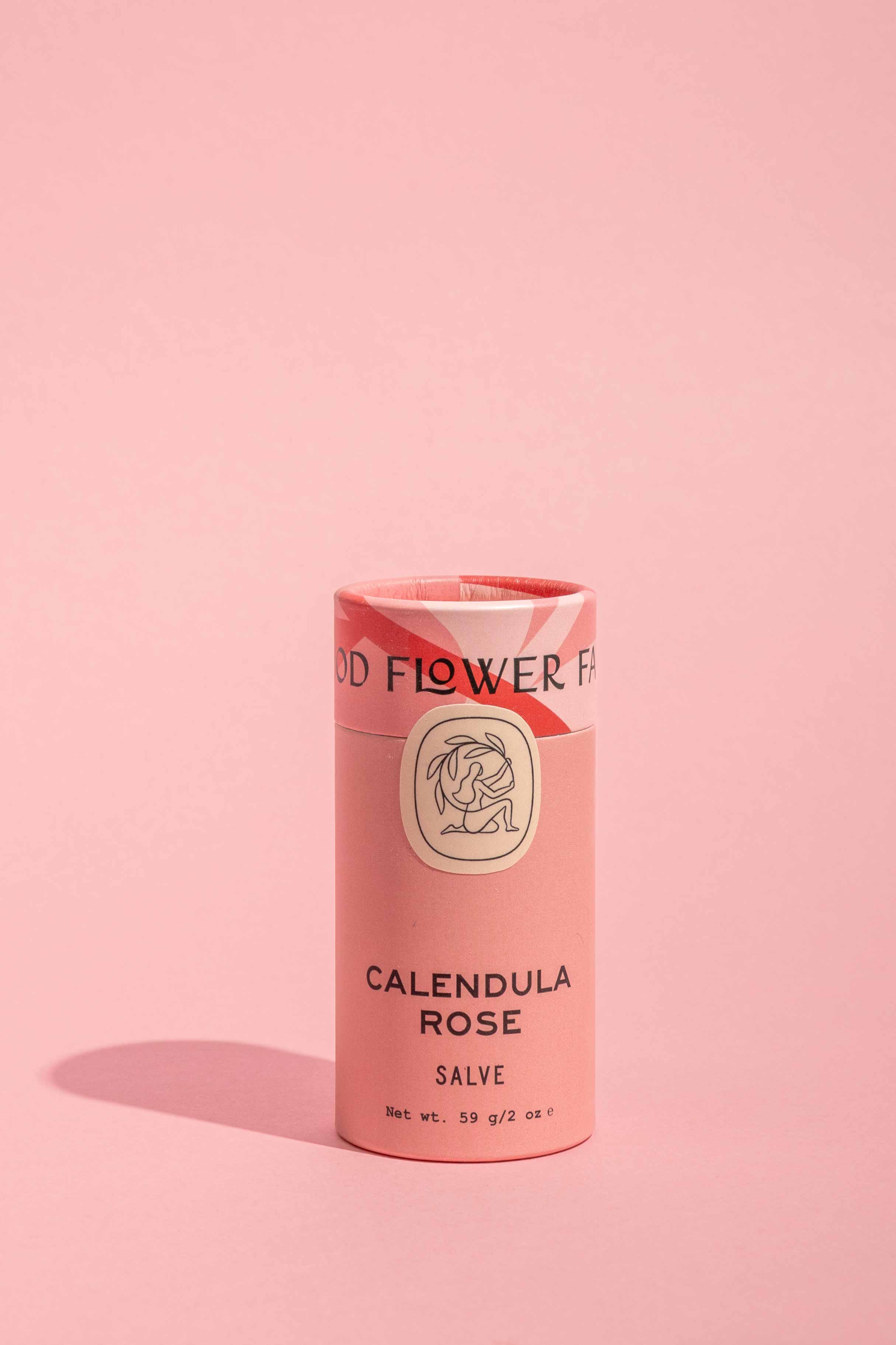 A pink cylindrical container of Good Flower Farm Calendula Rose Salve stands upright on a matching background, featuring simple black text, a floral illustration, and eco-friendly packaging.
