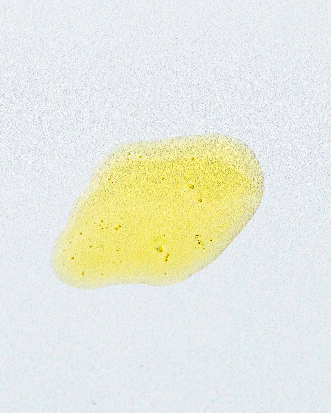 A small, irregularly shaped yellow liquid stain with bubbles—Matriarch Facial Serum by Good Flower Farm, enriched with helichrysum—rests on a white surface.