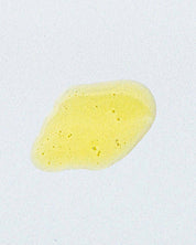 A small, irregularly shaped yellow liquid stain with bubbles—Matriarch Facial Serum by Good Flower Farm, enriched with helichrysum—rests on a white surface.