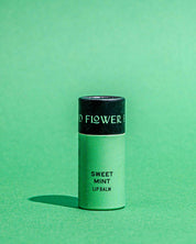 A green tube of Good Flower Farms TESTER — Sweet Mint Organic Lip Balm stands upright on a green surface, casting a shadow to the left. The moisturizing tube features a black cap with partial white lettering.