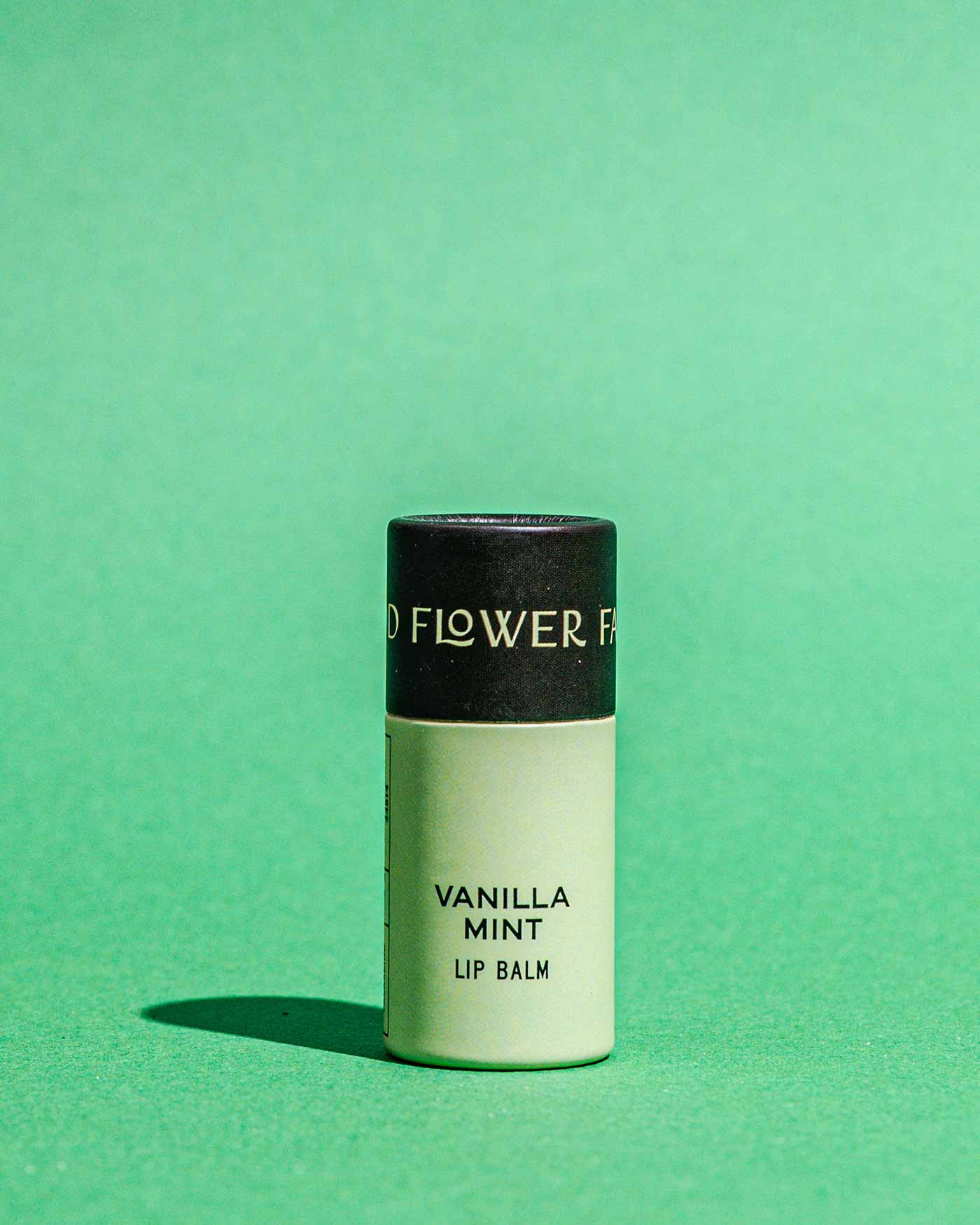 A tube of Good Flower Farms TESTER — Vanilla Mint Organic Lip Balm, featuring a black cap and light green body, stands upright on a green surface with a matching green background.