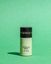 A tube of Good Flower Farms TESTER — Vanilla Mint Organic Lip Balm, featuring a black cap and light green body, stands upright on a green surface with a matching green background.