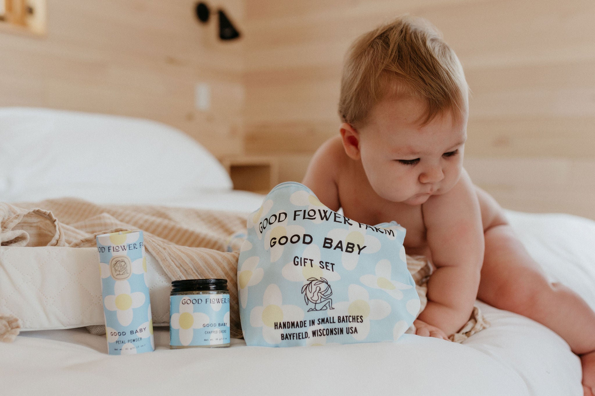 A baby on a bed reaches for the Good Flower Farm Good Baby Gift Set, which includes botanical skincare pouches, herbal baby powder in a jar, and diaper balm in a tube with blue and yellow floral designs.