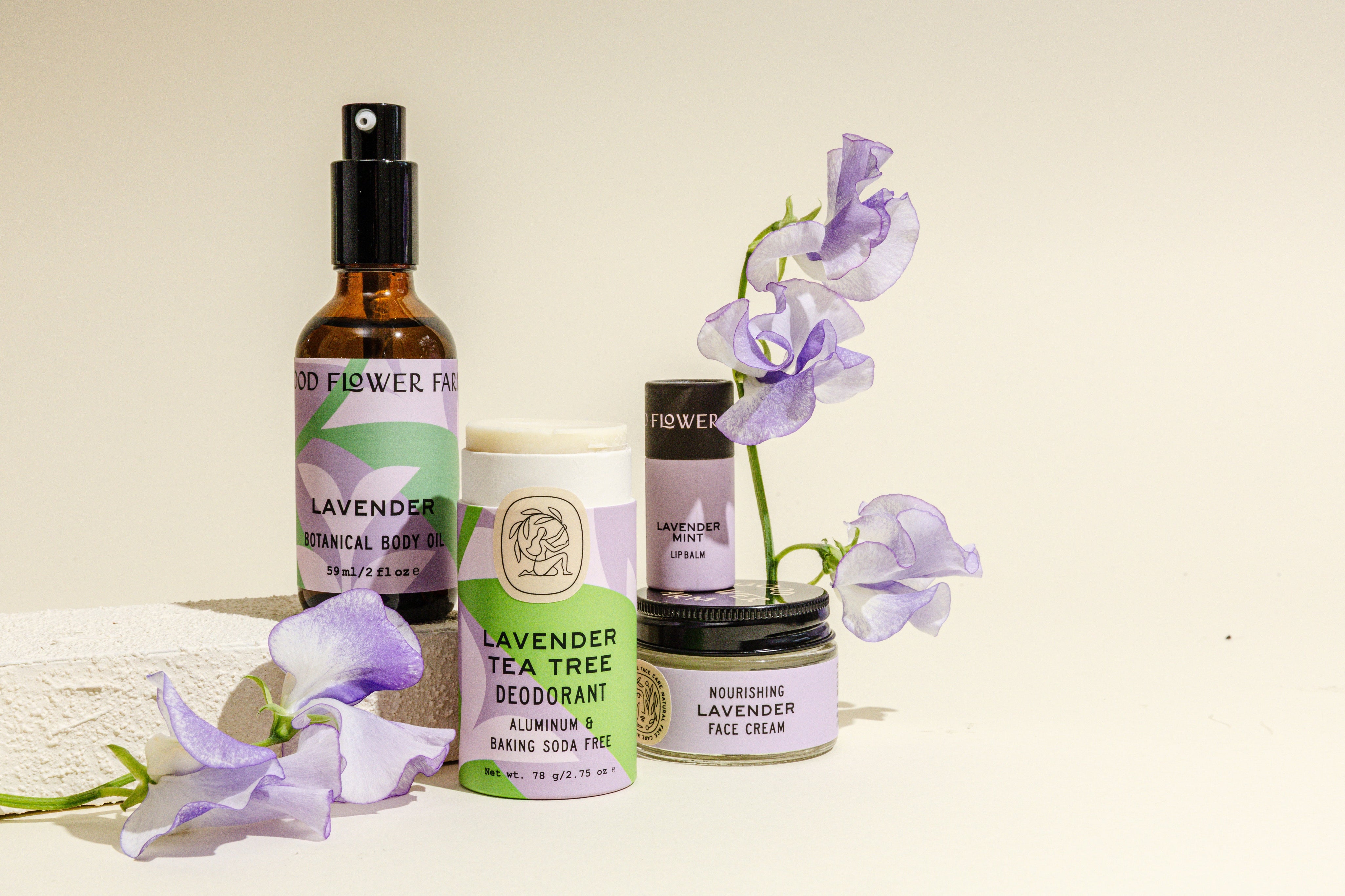 A collection of lavender skincare products, including body oil, deodorant, face cream, and lip balm, displayed with light purple flowers against a neutral background.