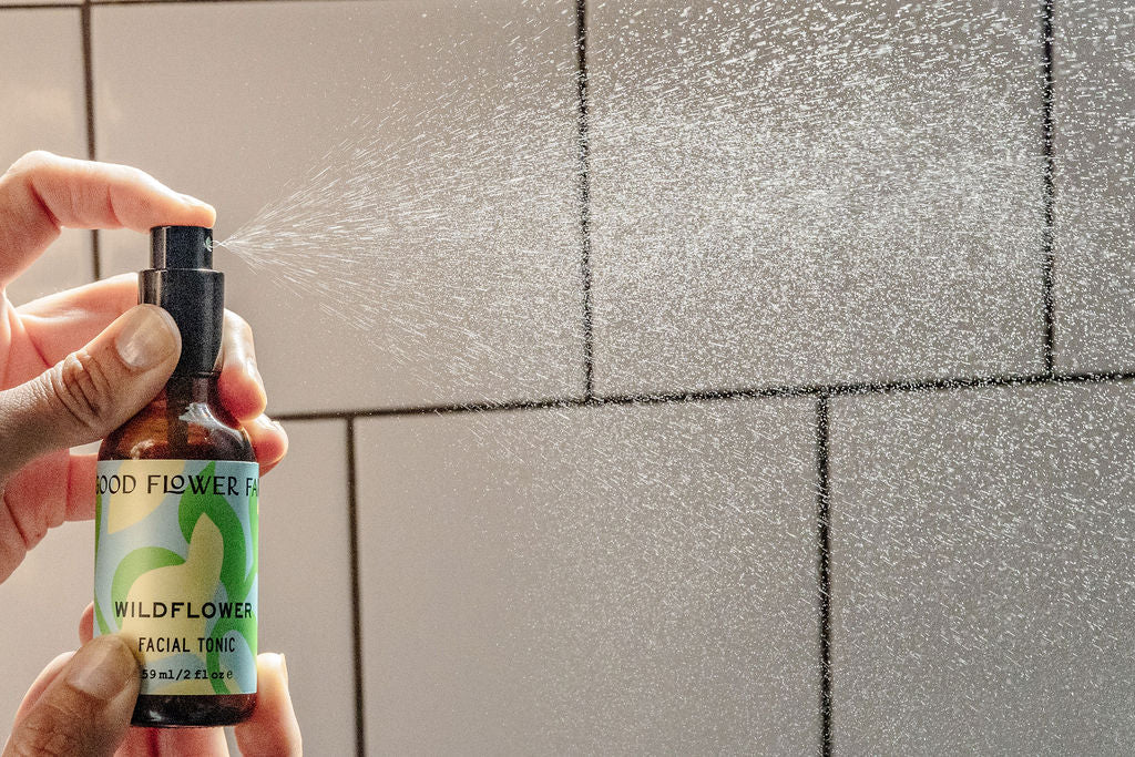 A hand sprays a mist of Wildflower Facial Tonic from a small brown bottle against a tiled wall, creating a fine spray in the air.