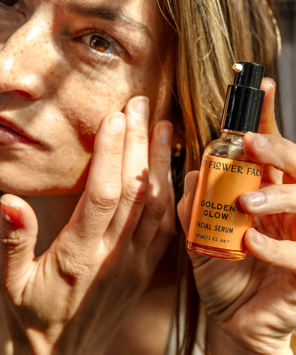 A woman applies facial serum to her cheek while holding a bottle labeled Golden Glow Facial Serum in bright sunlight, highlighting her skin and the product.