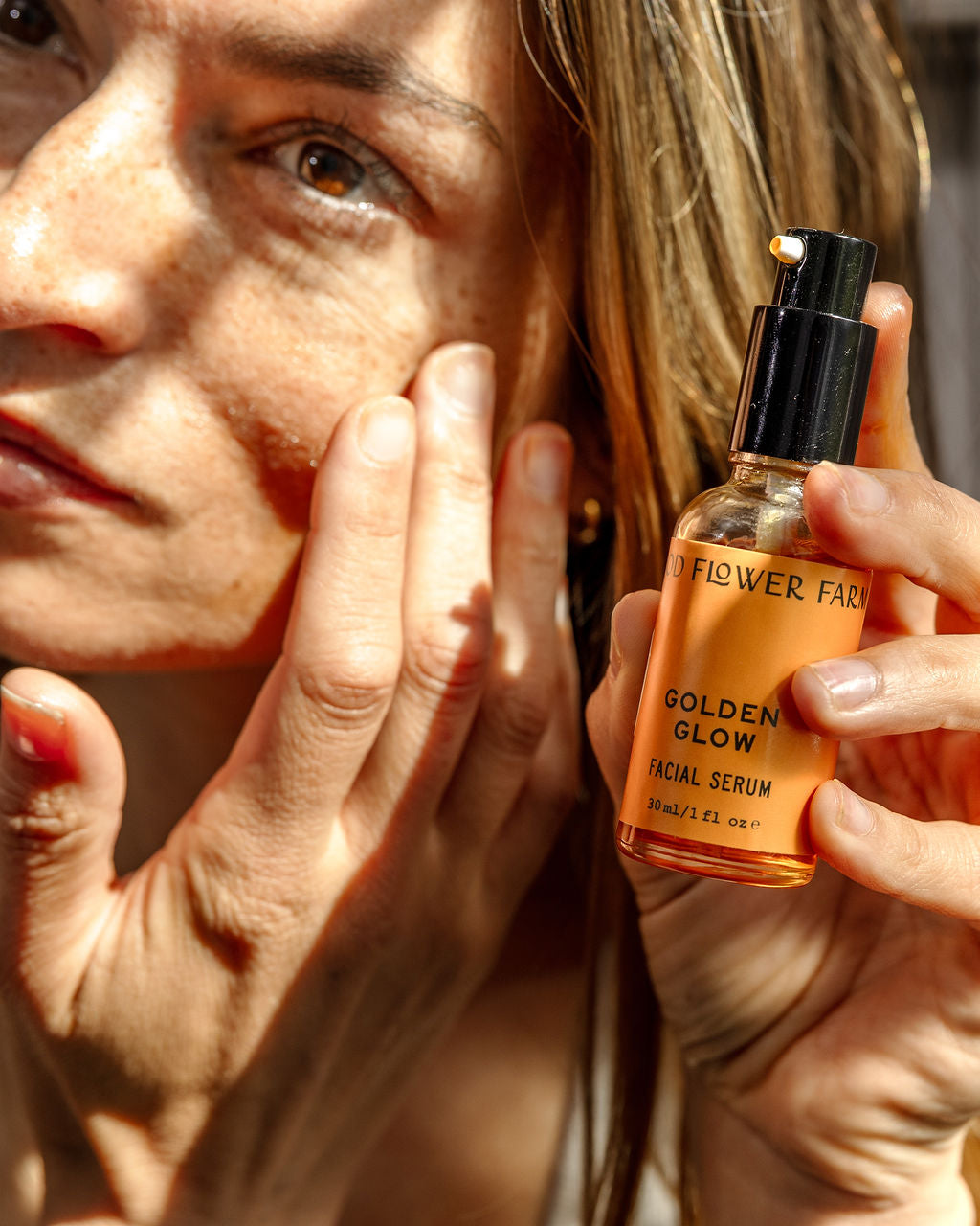 A woman applies facial serum to her cheek while holding a bottle labeled Golden Glow Facial Serum in bright sunlight, highlighting her skin and the product.