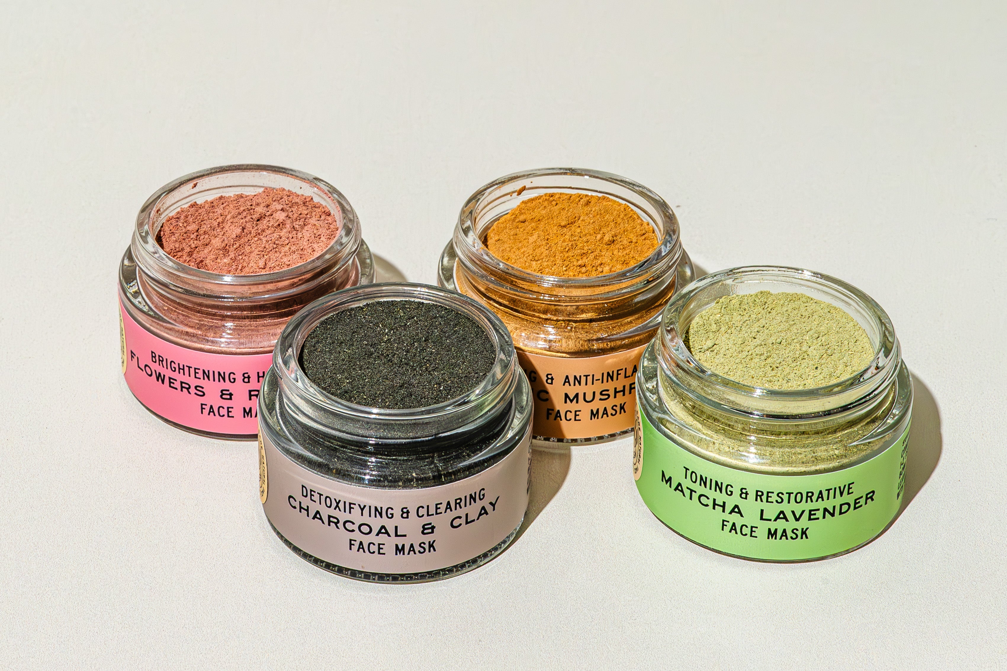 Four jars of powdered face masks are arranged on a light surface. Each jar contains a different colored powder and is labeled with its type and purpose, such as charcoal, mushroom, and matcha lavender.