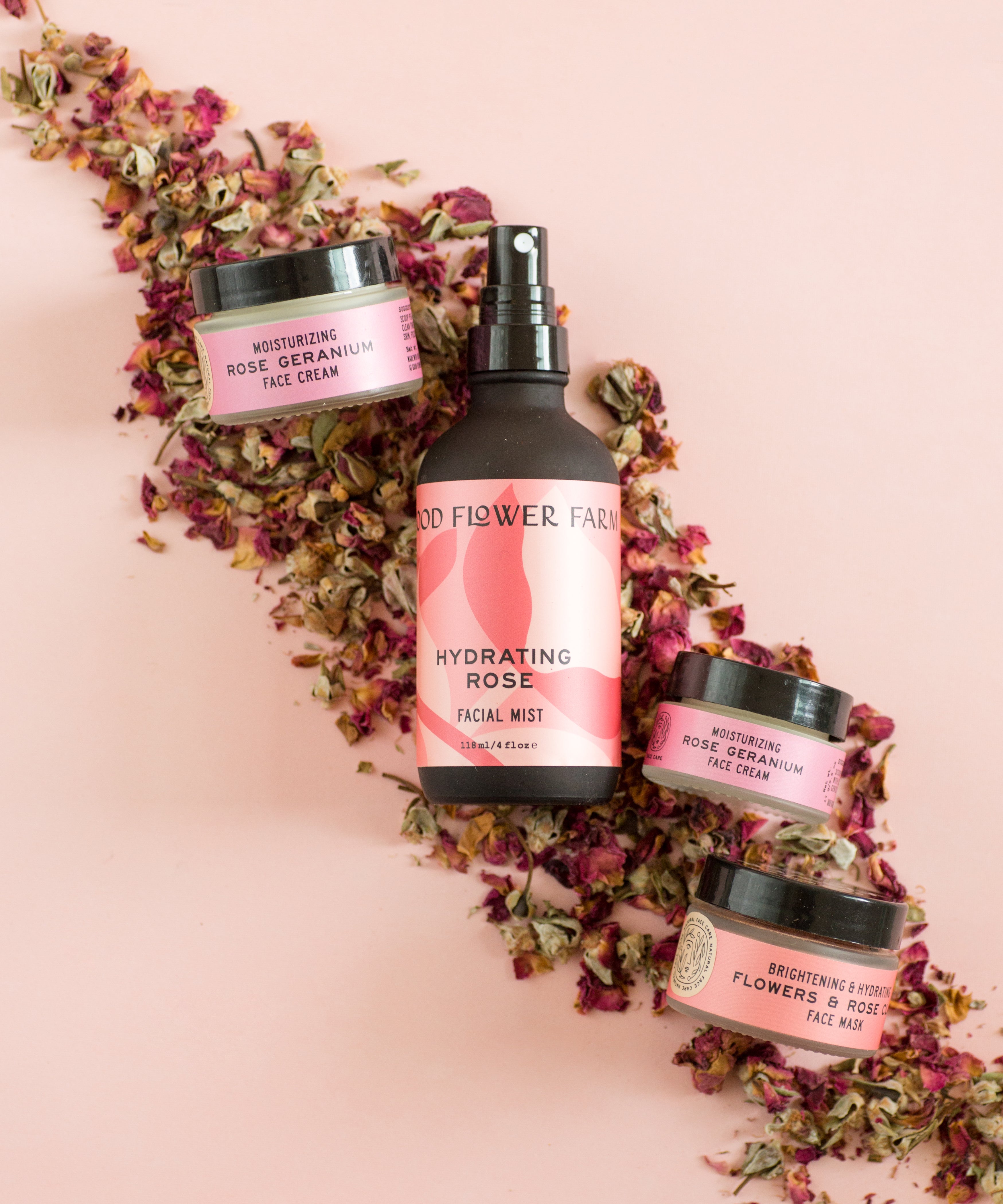 Four pink skincare products are arranged on dried rose petals against a pink background. A facial mist bottle is centered, surrounded by three small cream jars.