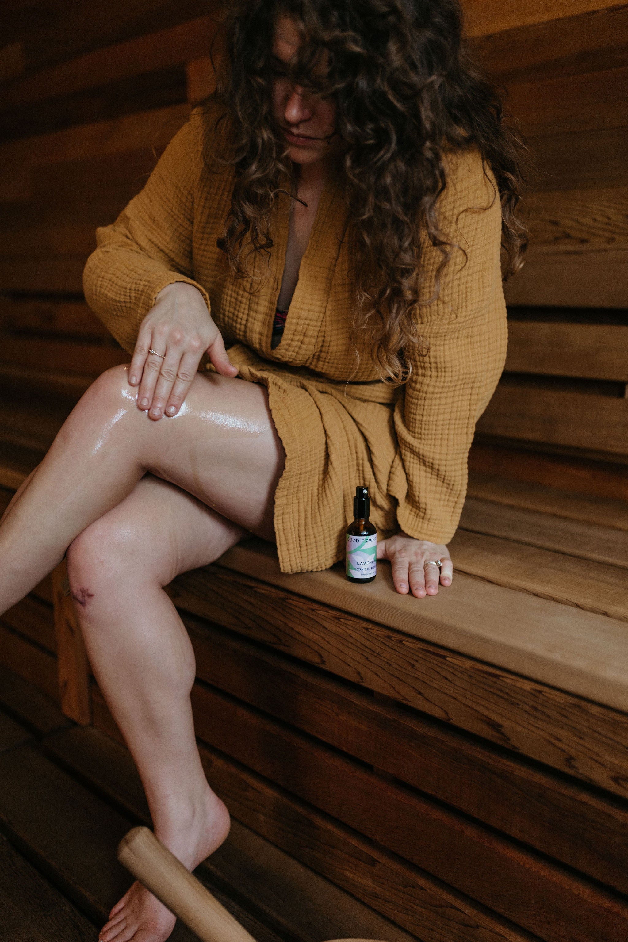 A woman in a mustard robe sits on a wooden bench, applying oil to her leg. A small bottle of oil is placed beside her on the bench.