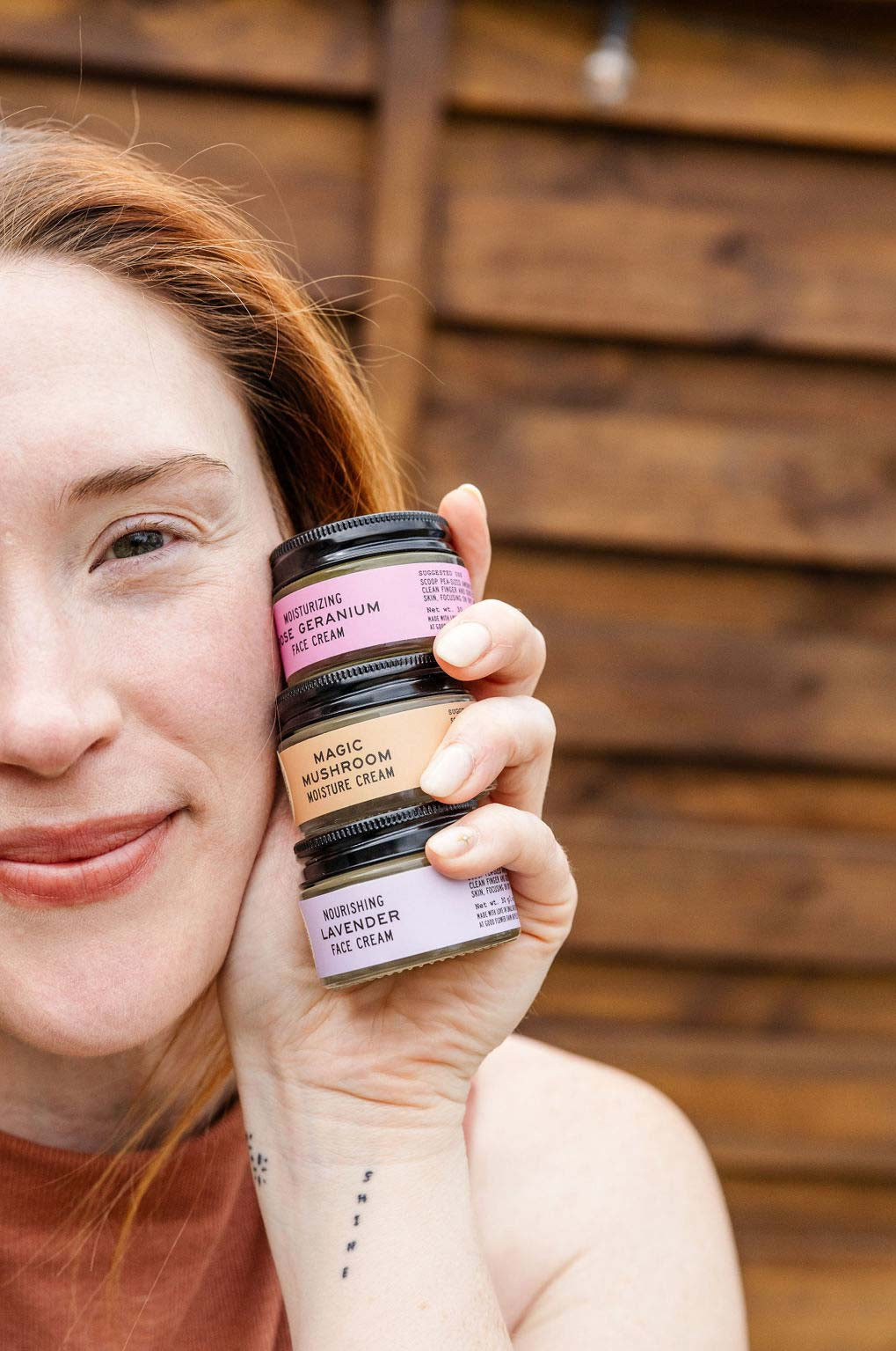 A woman with fair skin and auburn hair smiles, holding three small jars of face cream stacked by her cheek. The jars are labeled rose geranium, magic mushroom, and lavender. Wooden wall in background.