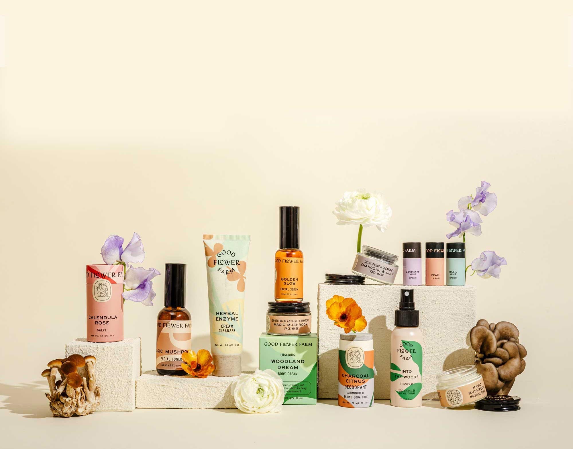 A collection of skincare products arranged on white blocks with colorful flowers and mushrooms, set against a pale background. The products have earthy, pastel packaging and include bottles, jars, and tubes.