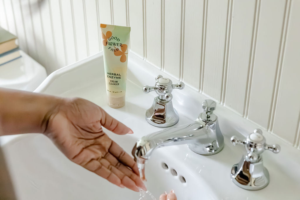 A person holds their hands under running water at a white sink, with a tube of herbal enzyme clay cleanser placed on the back of the sink.