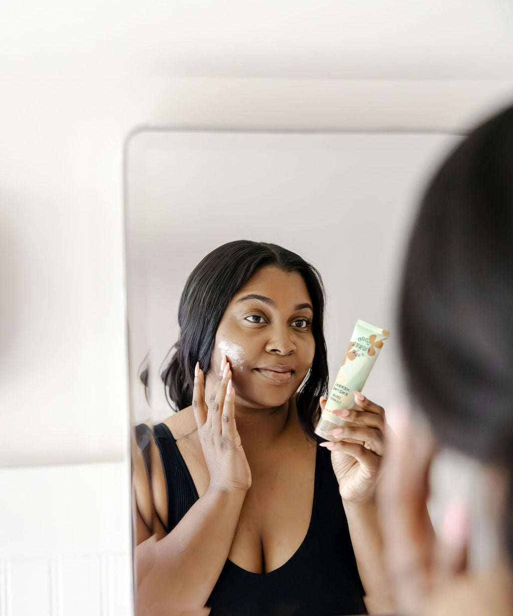 A woman applies cream to her face while looking in a mirror, holding a tube of skincare product in her hand and smiling gently, wearing a black top.