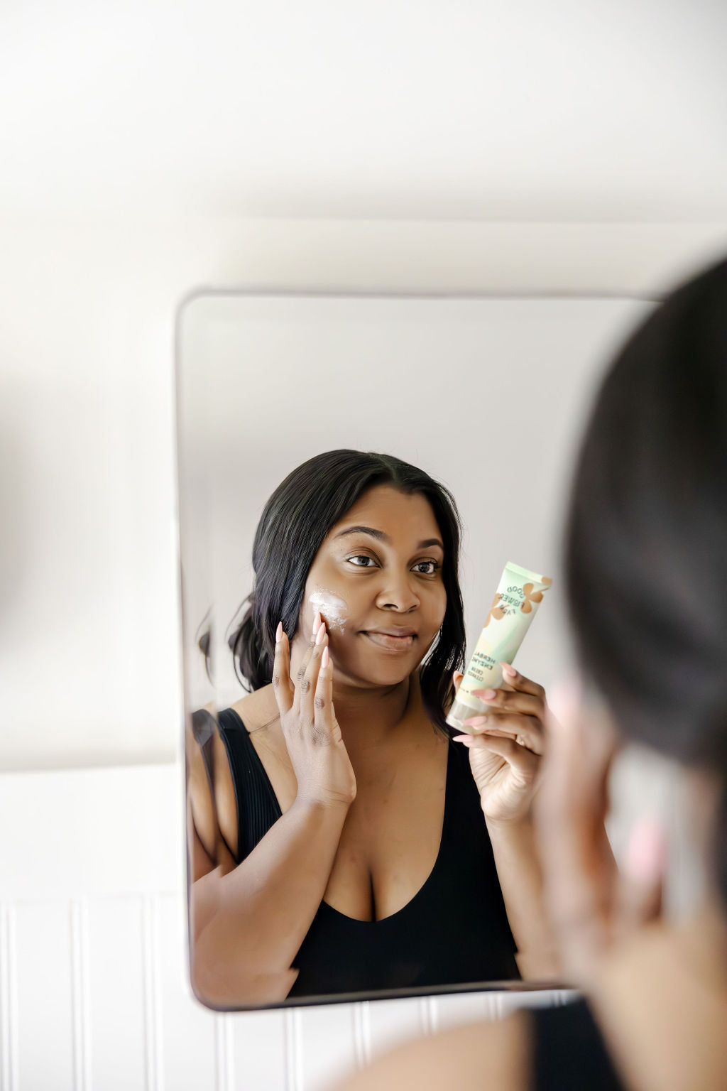 A woman applies cream to her face while looking in a mirror, holding a tube of skincare product in her hand and smiling gently, wearing a black top.