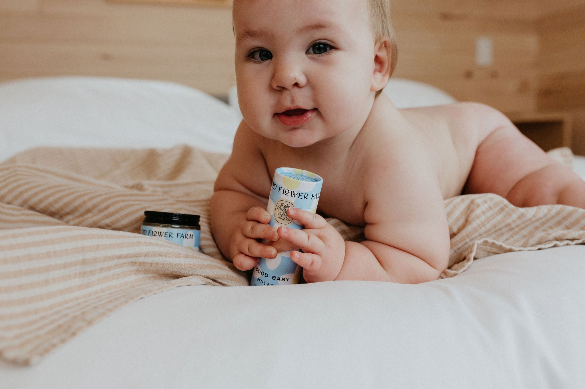 A baby lies on a bed holding a skincare product tube, with an open container nearby. The baby looks at the camera, surrounded by soft bedding in a cozy, wooden room.