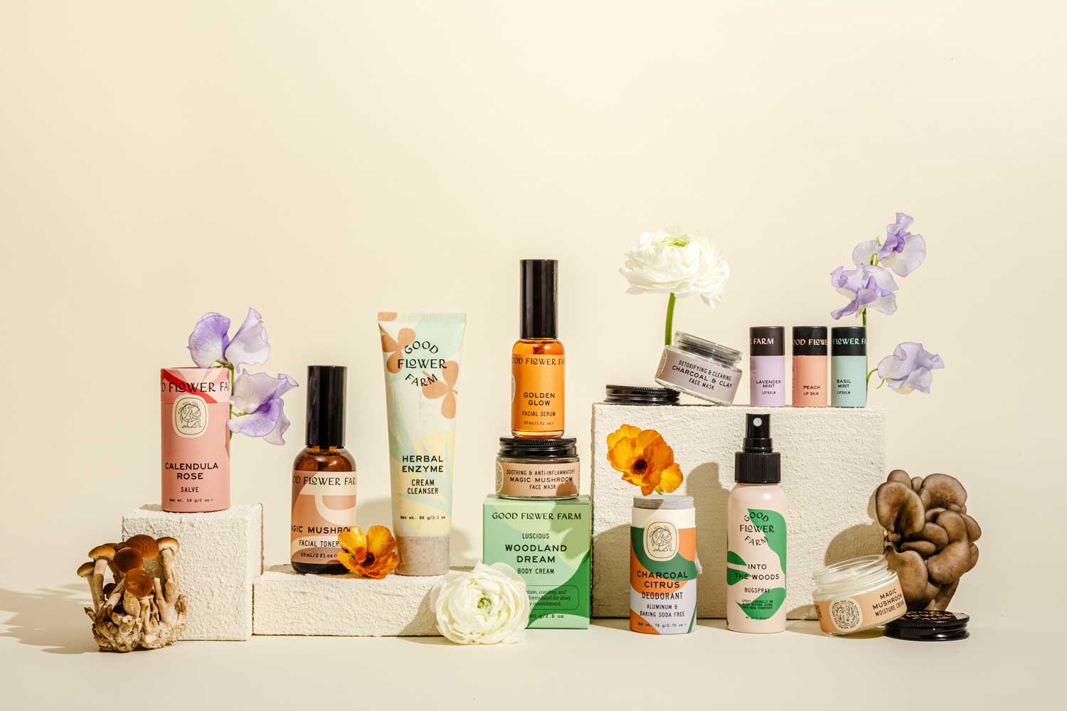 A collection of skincare and wellness products are arranged on white blocks, surrounded by fresh flowers and mushrooms, against a light beige background.