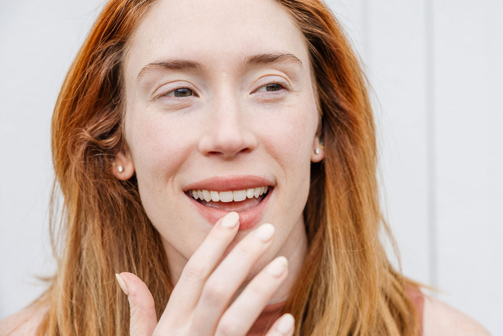 A woman with long reddish hair smiles, showing her teeth, while gently touching her chin with her index finger. She is wearing a sleeveless brown top.