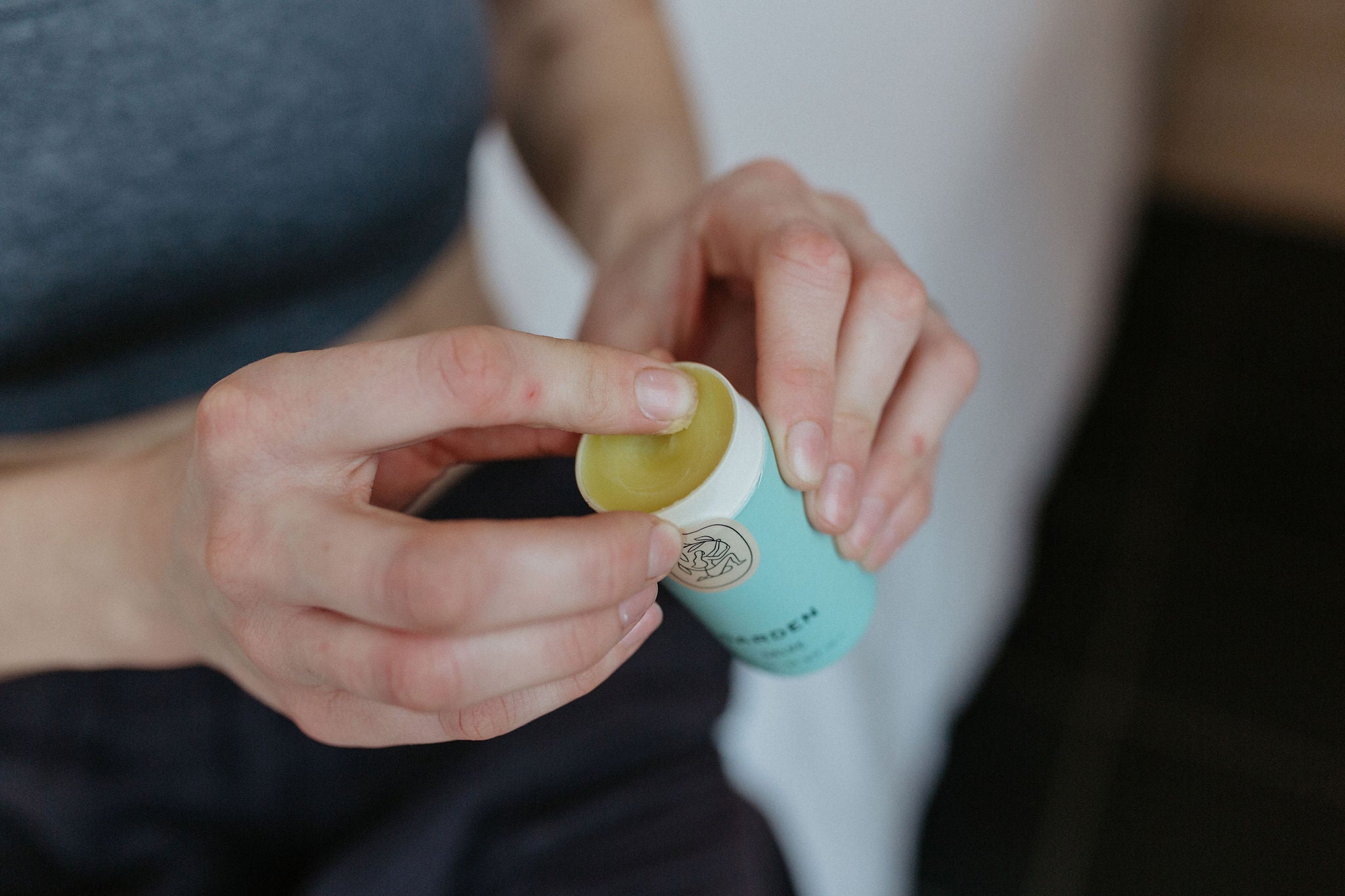 A person in a gray top uses their finger to scoop a yellow balm from a small, light blue container. The focus is on their hands and the balm.
