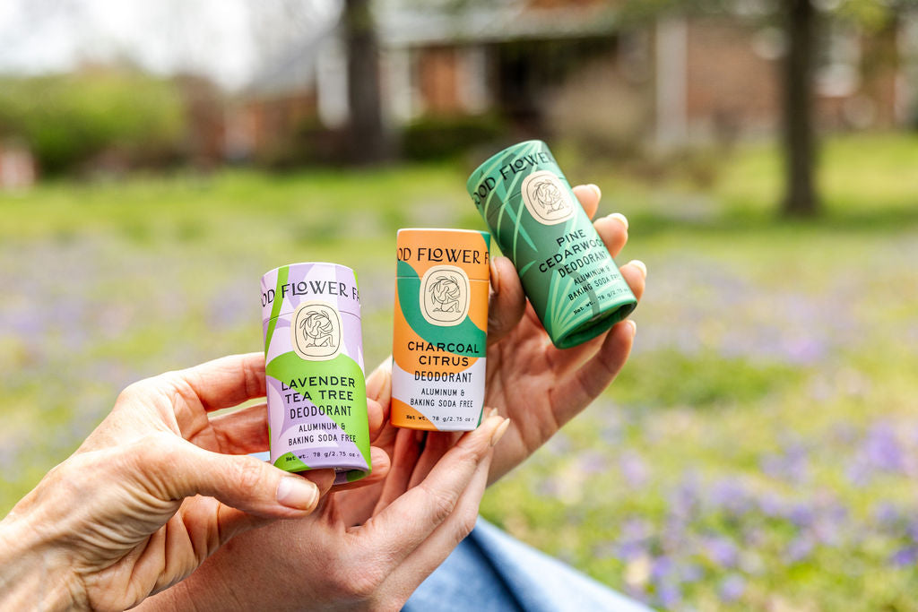 Two hands hold three natural deodorant sticks labeled Lavender Tea Tree, Charcoal Citrus, and Pine Cedarwood, with a grassy, flower-filled yard blurred in the background.