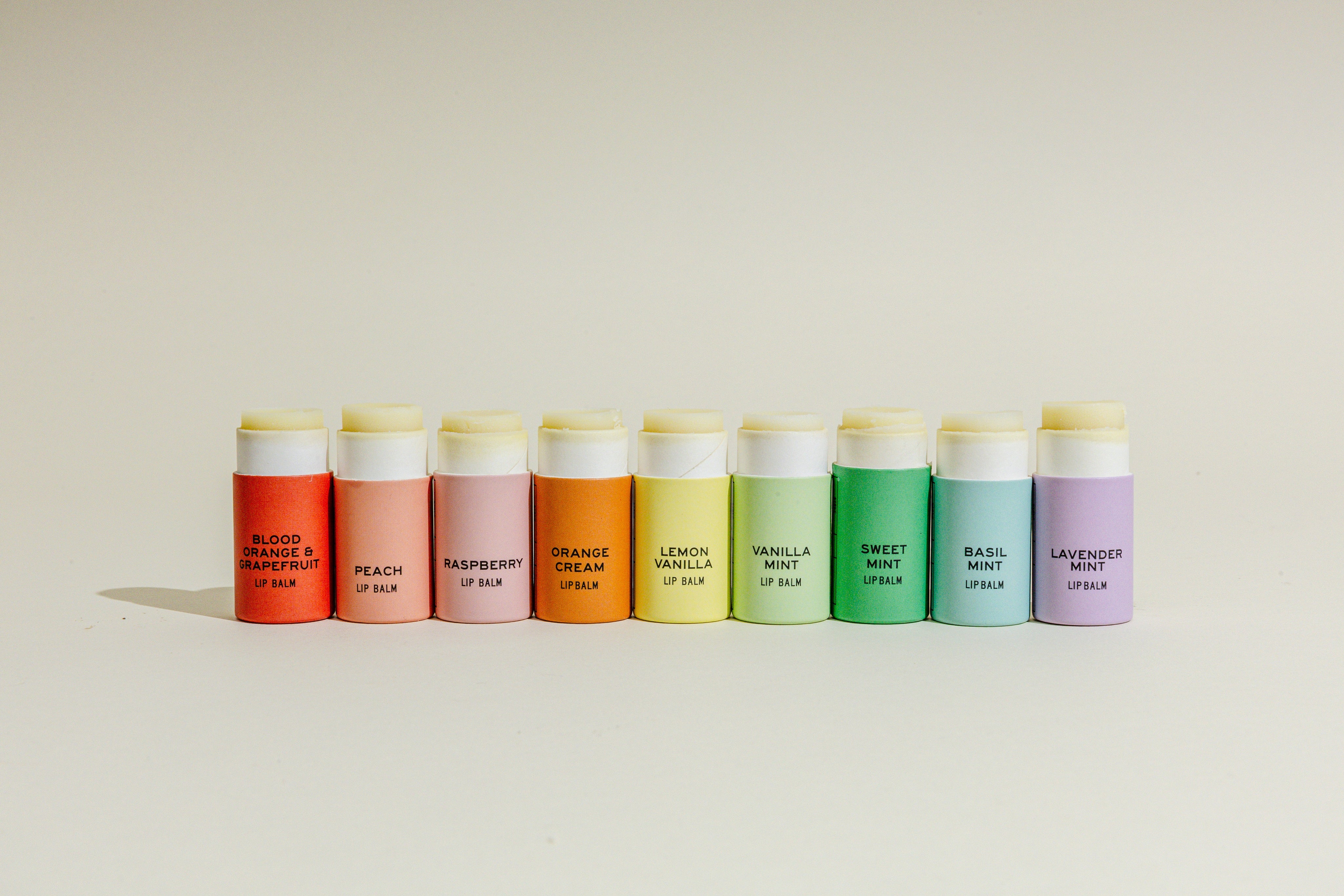 Nine colorful lip balm tubes in a row, each labeled with a different flavor, including blood orange, peach, raspberry, orange, lemon, vanilla, mint, basil mint, and lavender mint.