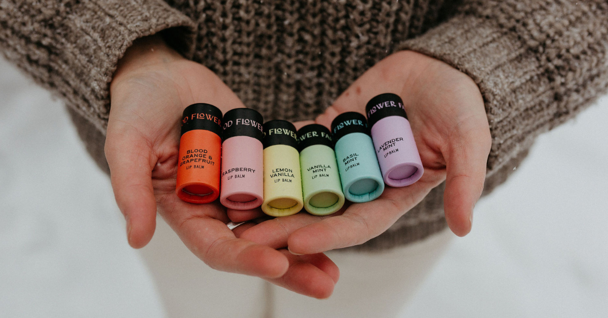 A person in a brown sweater holds six colorful lip balm tubes labeled with different scents, including blood orange, raspberry, vanilla, mint, and lavender, in their open hands.