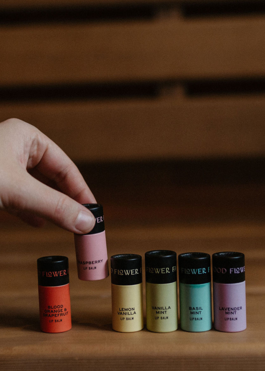 A hand picks a tube from the Good Flower Farm Rainbow Organic Lip Balm Set of 9, with nine colorful, moisturizing flavors displayed on a wooden surface against a blurred brown background.