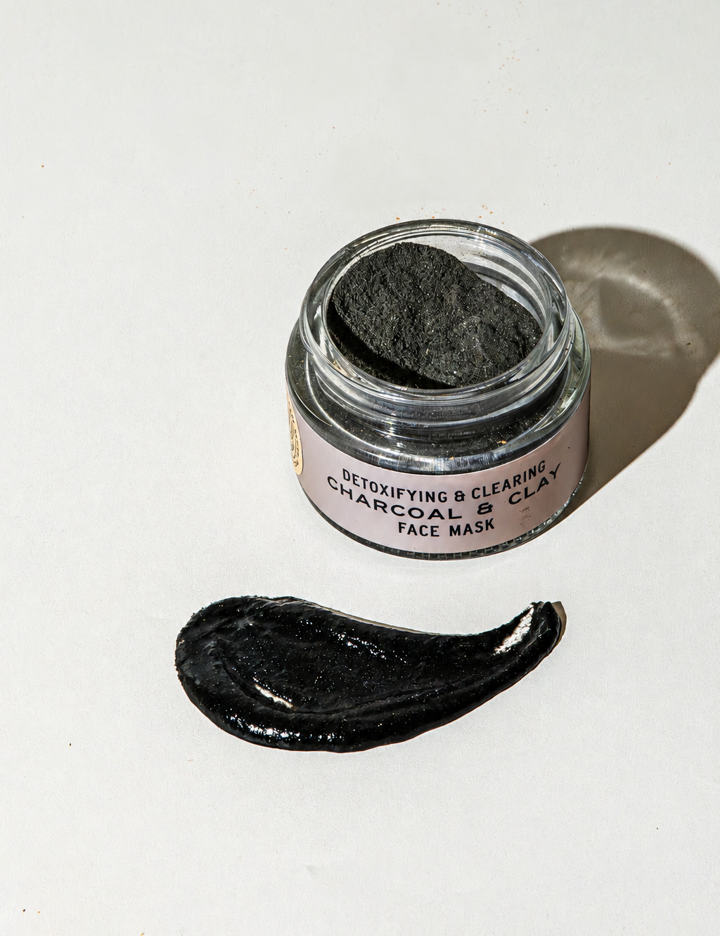 An open glass jar of Good Flower Farm Charcoal & Clay Botanical Face Mask next to a smear of the product on a white surface.