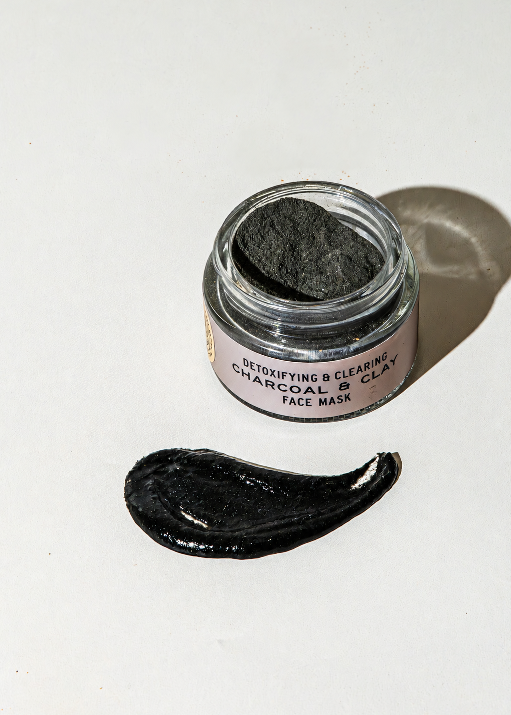 An open glass jar of Good Flower Farm Charcoal & Clay Botanical Face Mask next to a smear of the product on a white surface.