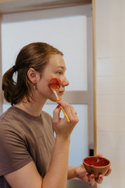 A woman applies the good-flower-farm Dry Skin Bundle: Save 15% facial mask to her cheek with a brush, holding a small bowl of the product in her other hand. Focused on moisturizing her face, she stands indoors wearing a brown shirt, with her hair pulled back.