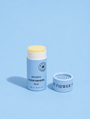 An open cylindrical blue container of Good Flower Farm's Everywhere Balm, unscented and labeled, sits on a light blue surface with its lid nearby. The yellow balm inside is slightly raised above the brim.