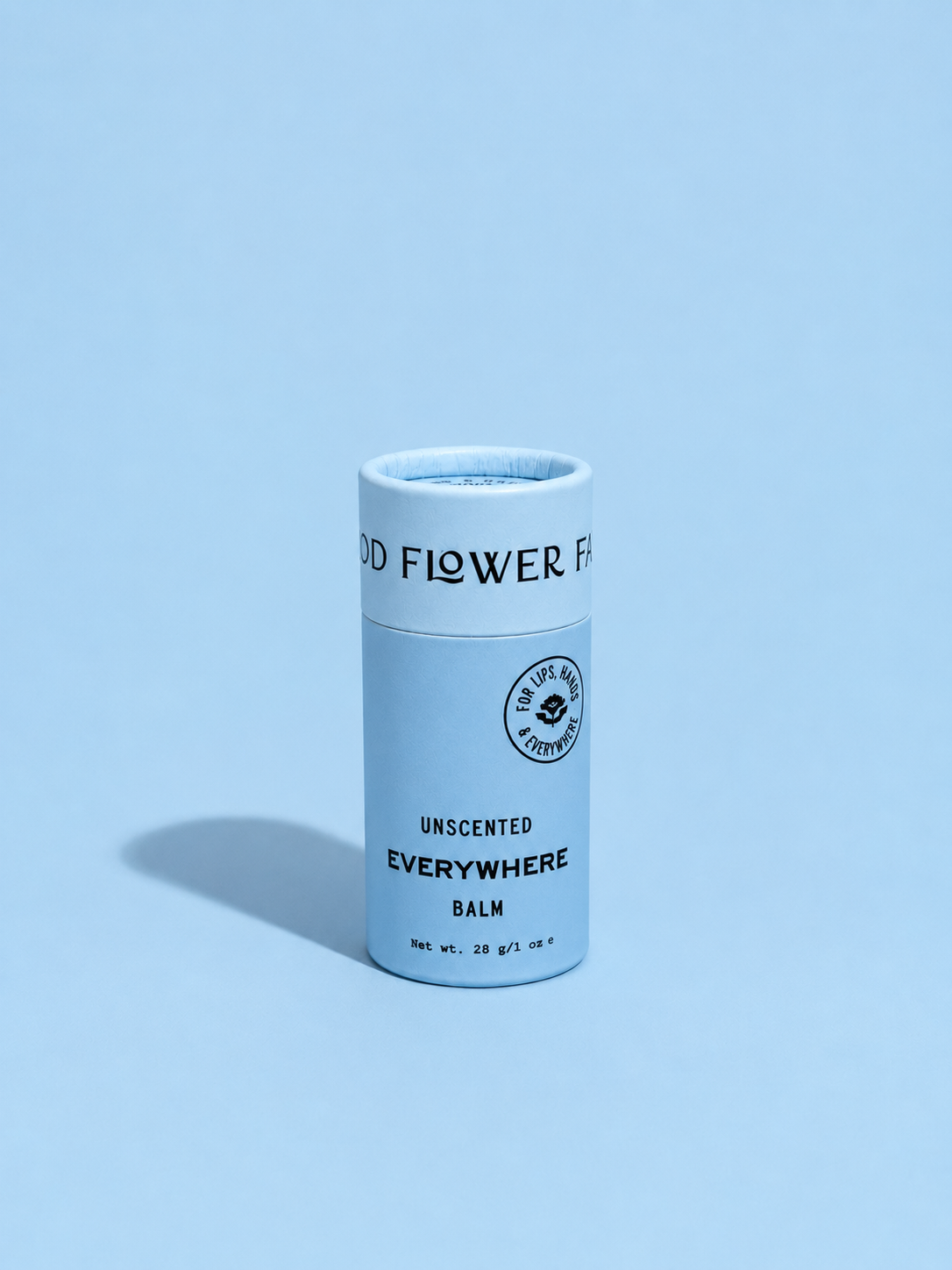 A cylindrical tube of Good Flower Farm’s Everywhere Balm stands upright on a light blue background with a soft shadow to the left. Its light blue packaging features black text.