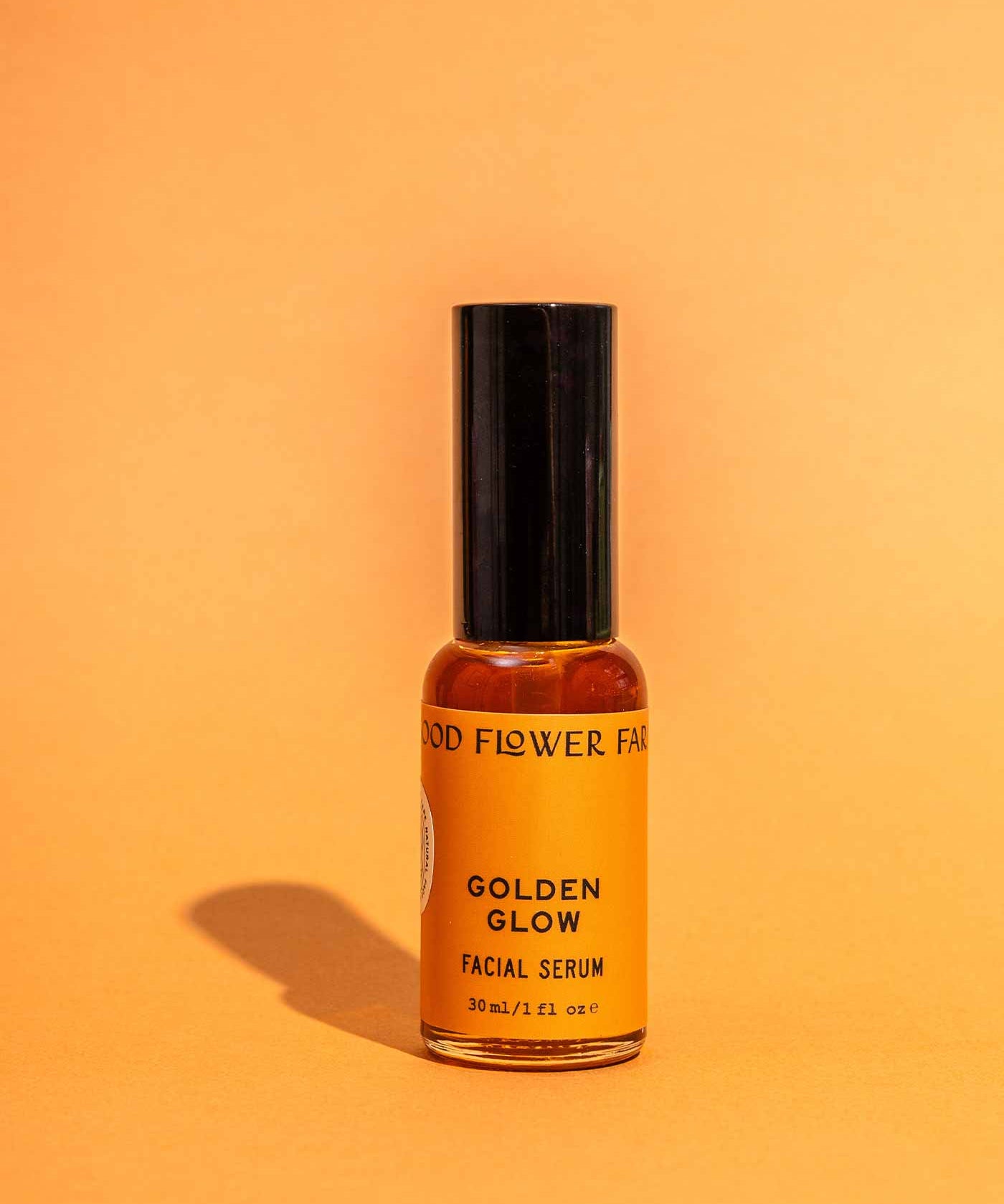 A small amber bottle labeled Golden Glow Facial Serum by Good Flower Farm stands upright with a black cap against an orange background, offering skin hydration and a natural glow.