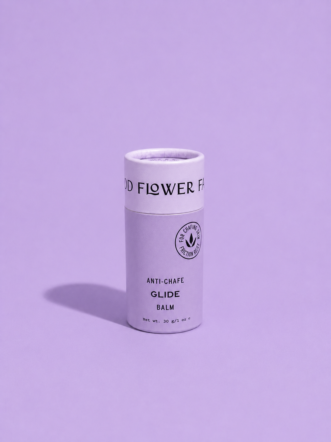 A cylindrical container of Good Flower Farm Glide Balm For Anti-Chafe stands upright on a purple surface with a matching background. Its light-colored label features black text and a small circular logo.