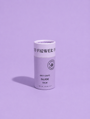A cylindrical container of Good Flower Farm Glide Balm For Anti-Chafe stands upright on a purple surface with a matching background. Its light-colored label features black text and a small circular logo.