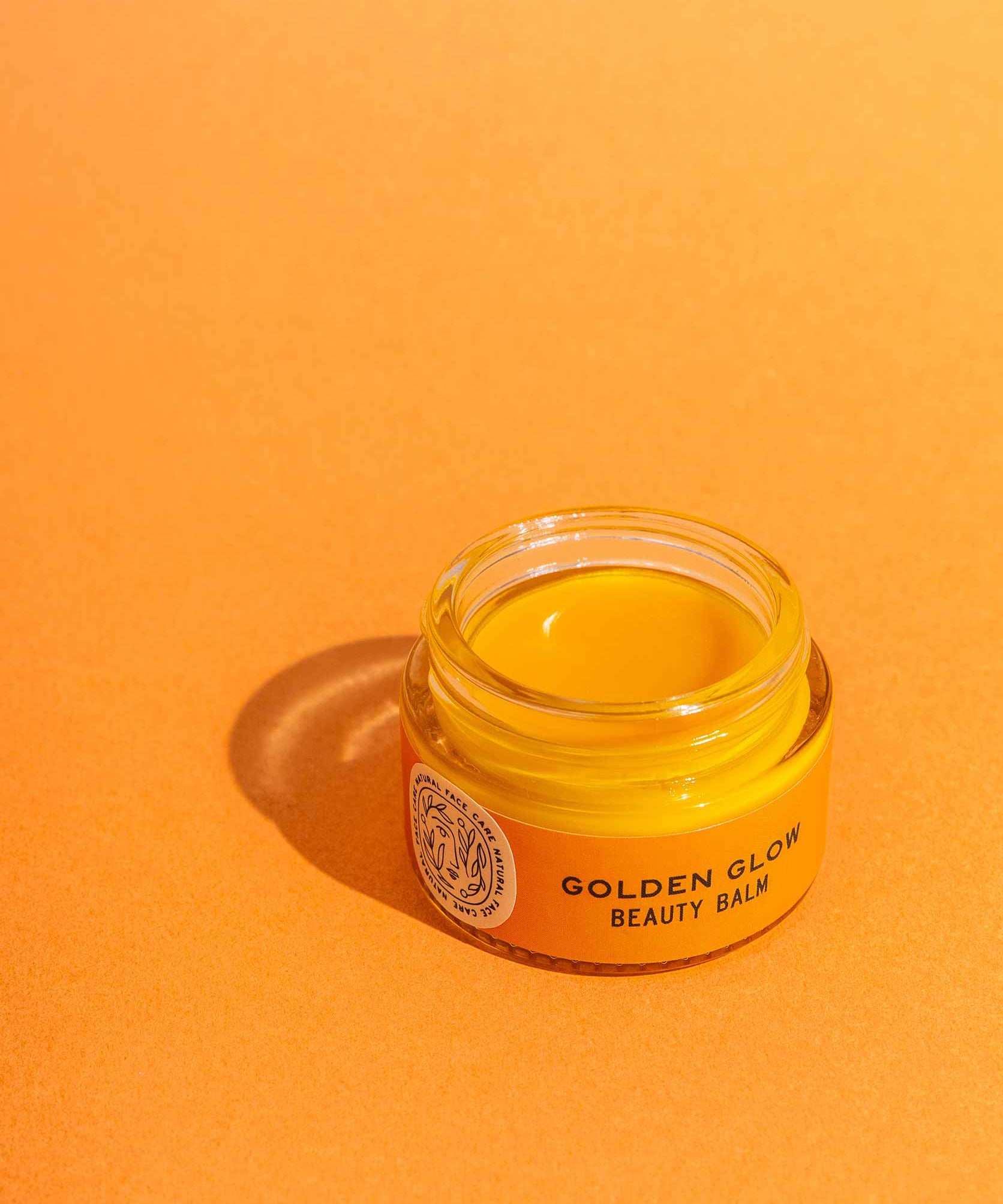 An open jar of Good Flower Farms Golden Glow Beauty Balm rests on an orange surface, its label and rich, nourishing contents illuminated by bright light—perfectly highlighting this skin repair balm.
