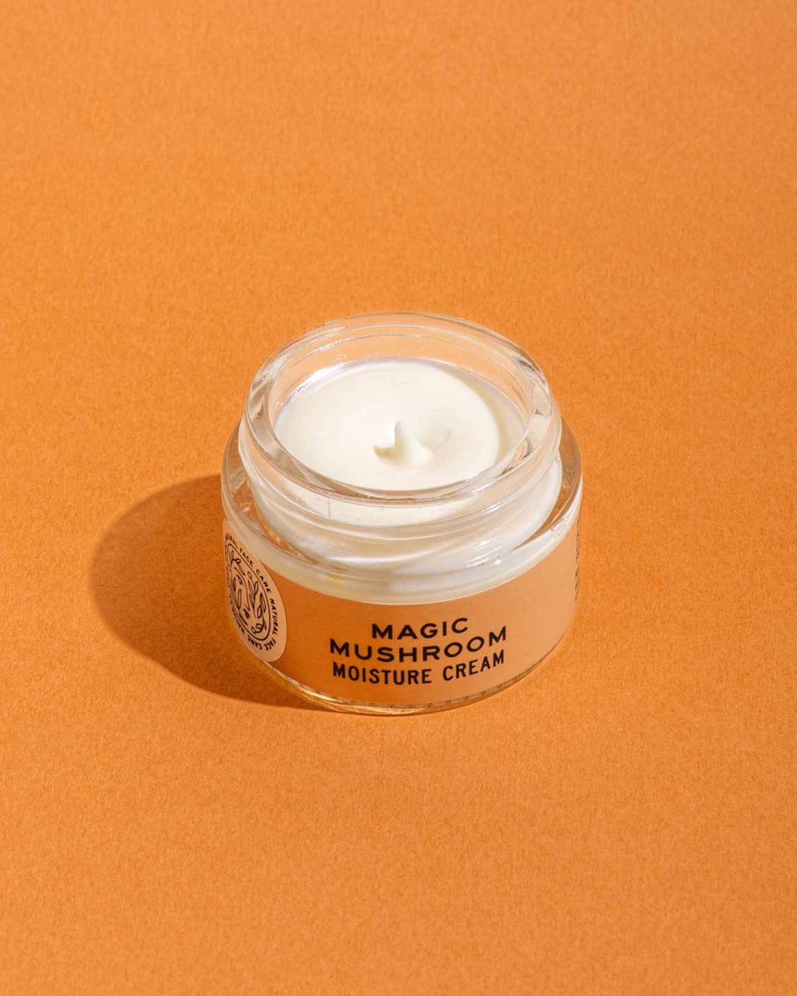 A small glass jar from good-flower-farm’s Dry Skin Bundle: Save 15% rests on an orange surface, filled with a creamy white moisturizer—ideal for facial hydration and soothing dry skin.