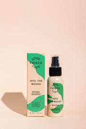 A bottle and box of Good Flower Farm’s Into the Woods Natural Bugspray, featuring green and beige packaging on a light peach background—made with essential oils for natural insect repellent.