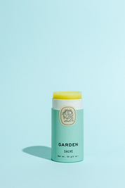 A light green tube of Good Flower Farm Garden Salve with a botanical logo and open lid reveals yellow balm inside, made from farm-grown herbs, displayed on a solid light blue background.