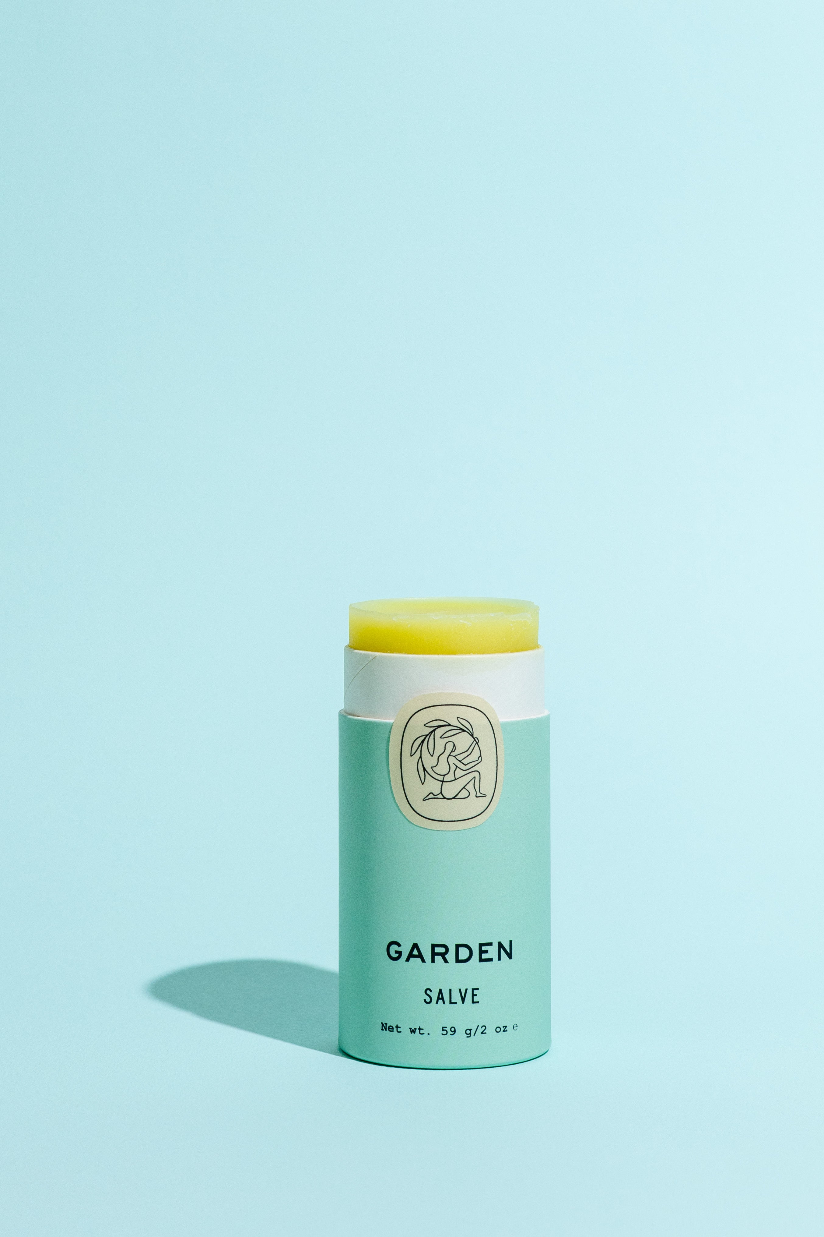 A light green tube of Good Flower Farm Garden Salve with a botanical logo and open lid reveals yellow balm inside, made from farm-grown herbs, displayed on a solid light blue background.